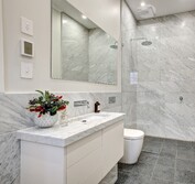 Interior view of ensuite showing stone vanity, wall hung toilet and open shower with glass screen. Ensuite with honed Carrara marble wall tiles and vanity with cut in sink. Honed black granite floor tiles.